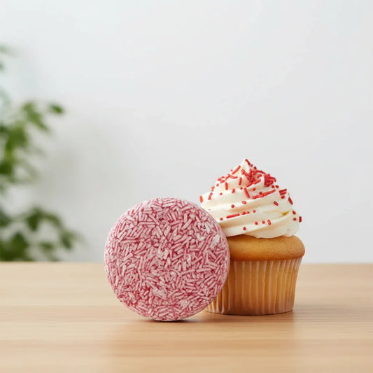 EPS bee hive shaped raspberry cupcake shampoo bar next to a vanilla cupcake with white frosting and red sprinkles on wooden surface